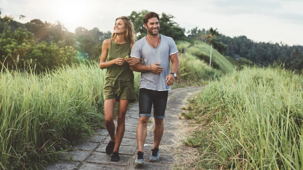 Texas- Couple walking through tall grass at sunset in a park on an old brink path. The woman is wearing a green romper, and the man is wearing a grey V-neck t-shirt and denim shorts with sneakers. New pet-friendly apartment homes for rent with private fenced backyards in Texas with pool, pickleball, dog park, gym, clubhouse and gated community. *No breed or weight restrictions. Yardly