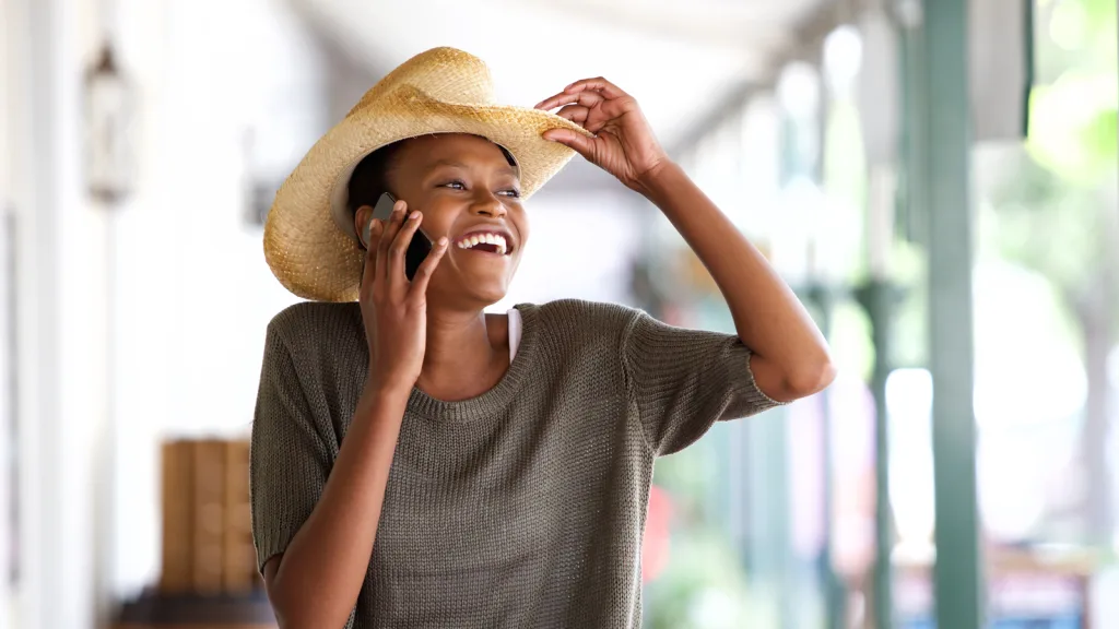 Texas-Woman holding her cowboy hat on her phone greeting someone by tipping her hat in the distance smiling and under an awning of a green and white barn. New pet-friendly apartment homes for rent with private fenced backyards in Texas with pool, pickleball, dog park, gym, clubhouse and gated community. *No breed or weight restrictions. Yardly
