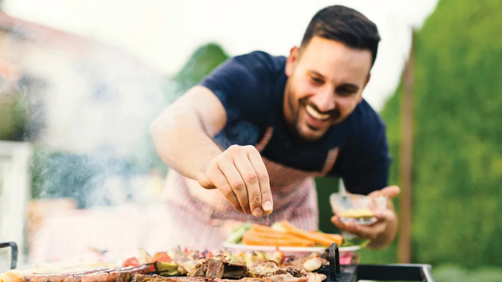 Man seasoning ribs on a grill smiling. He is wearing a plaid red and green apron. He holds his marinade seasoning bowl in his left hand and enthusiastically leans over sprinkling the seasonings with his right. New pet-friendly apartment homes for rent with private fenced backyards in Arizona, Texas, Florida, and North Carolina with pool, pickleball, dog park, gym, clubhouse and gated community. *No breed or weight restrictions. Yardly