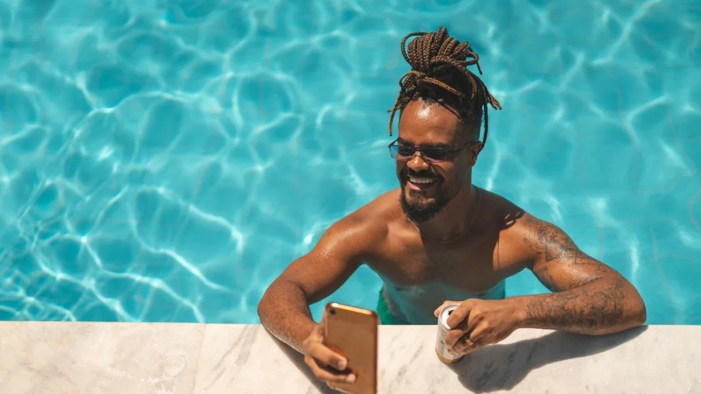 Man with goatee and indistinct tattoos on his left arm smiling in the pool on his phone and resting his arm and drink on the edge of the pool deck. New pet-friendly apartment homes for rent with private fenced backyards in Arizona, Texas, Florida, and North Carolina with pool, pickleball, dog park, gym, clubhouse and gated community. *No breed or weight restrictions. Yardly