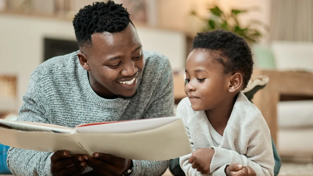 A father and son laying on their living room floor reading a book with their legs kicked up in the air behind them. the background setting has a soft-colored lamp, a plant, a couch, and a coffee table. New pet-friendly apartment homes for rent with private fenced backyards in Arizona, Texas, Florida, and North Carolina with pool, pickleball, dog park, gym, clubhouse and gated community. *No breed or weight restrictions. Yardly